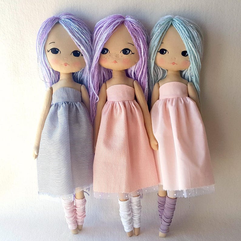 May include: Three handmade dolls with long, colorful yarn hair. Each doll is wearing a different colored dress. The doll on the left is wearing a gray and white striped dress, the doll in the middle is wearing a pink dress, and the doll on the right is wearing a light pink dress.