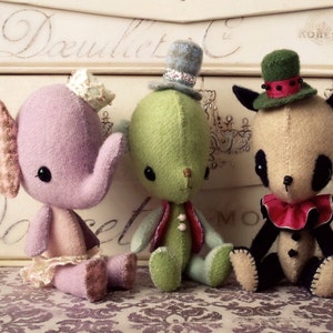 Può includere: Tre giocattoli di peluche fatti a mano: un elefante rosa con una corona, una creatura verde con un cappello blu e un panda marrone con un cappello verde con un bordo rosso.