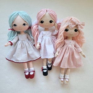 May include: Three handmade dolls with long hair. The doll on the left has blue hair and a striped dress. The doll in the middle has pink hair and a white dress. The doll on the right has pink hair and a pink and white striped dress.