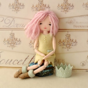 May include: A handmade doll with pink yarn hair, wearing a yellow dress and sitting on a small blue and green box. The doll is holding a small white crown in her hands.