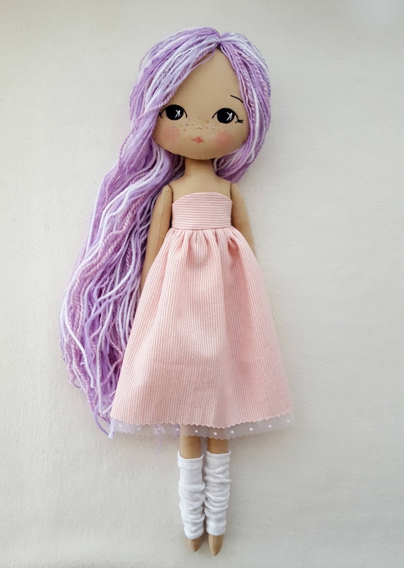 May include: A handmade doll with long, purple and white yarn hair, wearing a pink dress with a white lace trim and white socks.