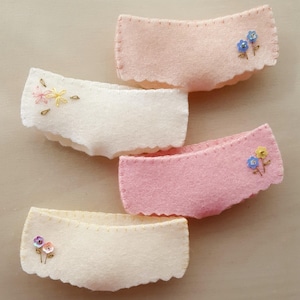 May include: Four felt fabric underwear with scalloped edges and embroidered flowers. The underwear are in shades of pink and cream.