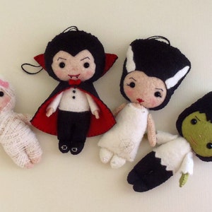 May include: Four felt Halloween ornaments, each depicting a different character. A mummy, a vampire, Frankenstein's monster, and a witch. The ornaments are all white with black and red details.