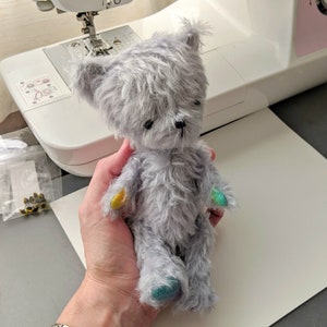 以下が含まれることがあります： 手のひらサイズのグレーのふわふわのテディベア。足にカラフルなアクセントが施されています。ベアは人の手に持たれています。