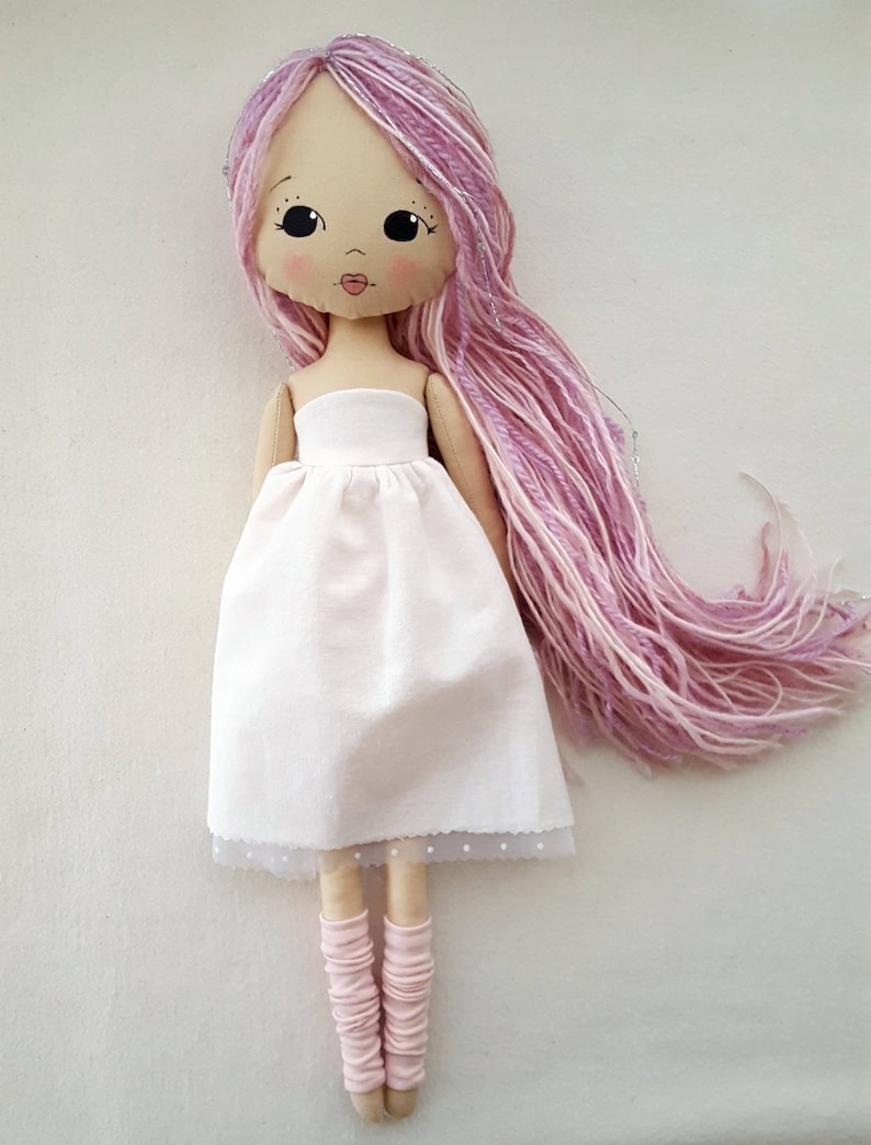 May include: A handmade doll with long pink and white yarn hair, wearing a white dress with a lace trim and pink leg warmers.