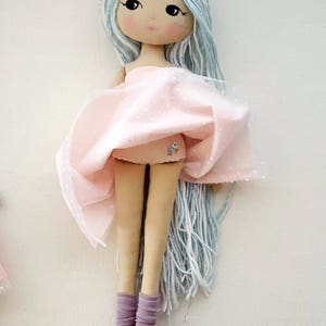 May include: A handmade doll with long, light blue yarn hair, wearing a pink tulle skirt with white polka dots and purple leg warmers.