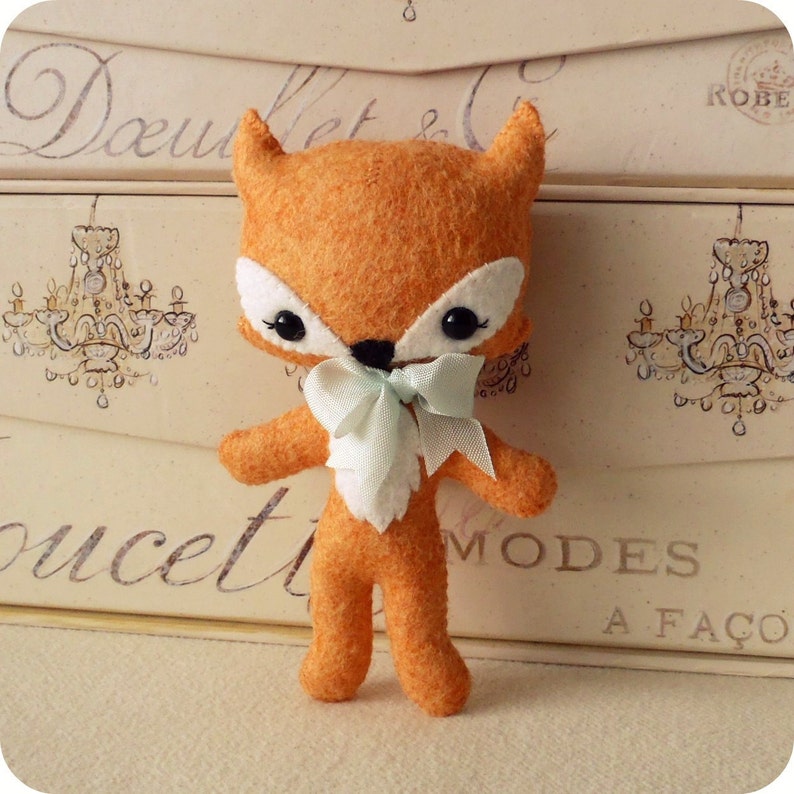 May include: A small orange felt fox toy with a white belly and a light blue bow. The fox has black eyes and a black nose.