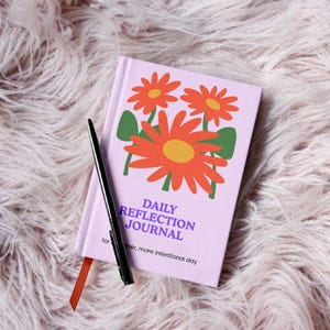 May include: A lavender Daily Reflection Journal with orange flower illustrations and the text "Daily Reflection Journal". A black pen rests on the journal with a red ribbon bookmark.