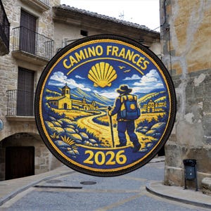 May include: Round patch with the words "CAMINO FRANCES 2026". The design shows a hiker with a backpack and walking stick on a path, a yellow scallop shell, and a landscape of buildings and mountains.