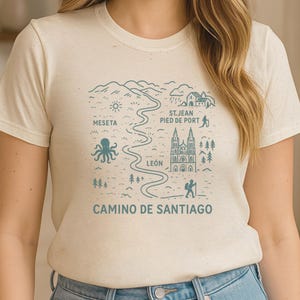 以下が含まれることがあります： クリーム色のTシャツ。ターコイズブルーのサンティアゴ巡礼路の地図デザイン。ランドマーク、テキスト、タコのイラストが含まれています。テキストには「MESETA」、「LEÓN」、「ST. JEAN PIED DE PORT」、「CAMINO DE SANTIAGO」が含まれています。