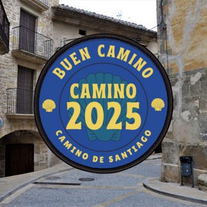 Puede incluir: Un parche redondo azul con las palabras "BUEN CAMINO CAMINO 2025 CAMINO DE SANTIAGO" en amarillo. El parche presenta un diseño de concha de vieira y está sobre un fondo de edificios de piedra.