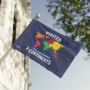 May include: A navy blue flag with the words "VISITED 7 CONTINENTS" in white. A colorful world map is in the center, with each continent highlighted in a different color. The flag is attached to a silver pole and is waving in the wind.