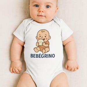 May include: White baby onesie with a cartoon illustration of a baby holding a shell. The illustration is in brown and beige tones. The text "BEBÉGRINO" is printed in navy blue below the illustration.