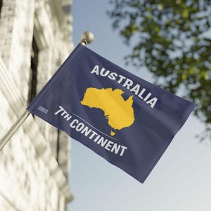 Op de afbeelding: Een marineblauwe vlag met de tekst "AUSTRALIA" en "7TH CONTINENT" in het wit. Een gele omtrek van Australië staat in het midden. De vlag is bevestigd aan een zilveren paal en wappert in de wind.