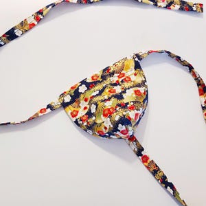 May include: A Japanese-style eye patch with a floral pattern. The fabric features a navy blue background with red, white, and gold floral designs. The straps are also patterned with similar floral motifs.