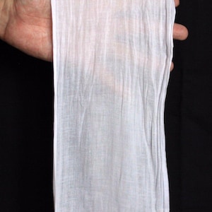 Può includere: Un tessuto bianco e semitrasparente su sfondo nero. Il tessuto è finemente tessuto e leggermente stropicciato, con linee verticali. Il materiale appare leggero e delicato, potenzialmente per abbigliamento o decorazioni.