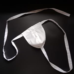 Könnte beinhalten: Ein weißer Sport-BH aus Stoff mit langen, dünnen Trägern. Der BH hat eine Tasche und Träger, die um die Taille und die Beine gebunden werden. Der Hintergrund ist schwarz.