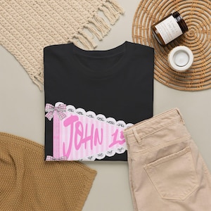 Könnte beinhalten: Ein schwarzes T-Shirt mit einer Grafik eines rosa-weiß gestreiften Wimpels, verziert mit weißer Spitzenborte und zwei kleinen grauen Schleifen mit Punkten. Der Text JOHN 15 ist in einer kräftigen rosa Schreibschrift auf dem Wimpel gedruckt.