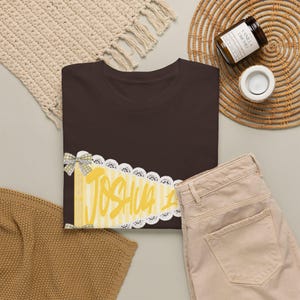 May include: A dark brown t-shirt featuring a yellow pennant graphic with white vertical stripes and a white lace border. The name JOSHUA is printed in a bold yellow script font across the centre of the pennant, accented by two small grey gingham bows.