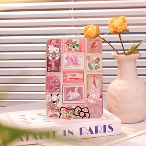May include: A decorative phone case featuring a collage of vintage postage stamps with floral and cartoon character designs. The case includes images of Hello Kitty, a swan, and other illustrations. The case is resting on a book titled "A Table in Paris".