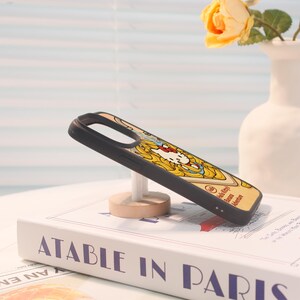 Może przedstawiać: Czarne etui na telefon z motywem Hello Kitty na ż&oacute;łtym tle, z napisem "Hello Kitty Banana Adventure". Etui jest prezentowane na drewnianym stojaku, spoczywającym na książce zatytułowanej "ATABLE IN PARIS".