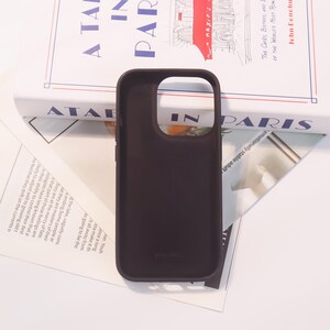 Może przedstawiać: Ciemnobrązowe etui na telefon z wycięciem na aparat i napisem "14 PRO" na dole. Etui leży na książce zatytułowanej "A Table in Paris".
