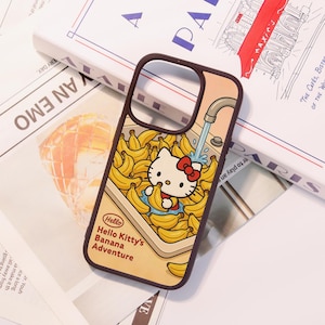 Może przedstawiać: Brązowe etui na telefon z kresk&oacute;wkową ilustracją Hello Kitty w zlewie pełnym banan&oacute;w. Etui zawiera napis "Hello Hello Kitty's Banana Adventure". Tło jest w jasnym kolorze brzoskwiniowym.