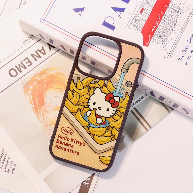 Może przedstawiać: Brązowe etui na telefon z kresk&oacute;wkową ilustracją Hello Kitty w zlewie pełnym banan&oacute;w. Etui zawiera napis "Hello Hello Kitty's Banana Adventure". Tło jest w jasnym kolorze brzoskwiniowym.