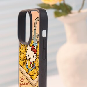 Może przedstawiać: Czarne etui na telefon z kresk&oacute;wkową ilustracją Hello Kitty w motywie banan&oacute;w. Projekt zawiera kran z bieżącą wodą i tekst "Hello Kitty's Bananas". Obudowa ma czarną obw&oacute;dkę.