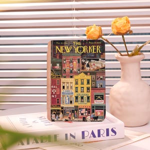 Puede incluir: Un bloque de madera decorativo con una colorida ilustración de una escena callejera de Nueva York, con el texto "THE NEW YORKER" y "Mar 1944". El bloque descansa sobre un libro titulado "ATABLE IN PARIS". Un jarrón con flores amarillas está en el fondo.