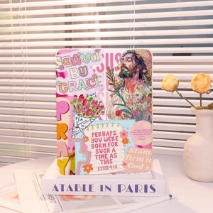 Peut inclure: Un livre décoratif avec le texte "Saved by Grace" et "Pray". Le livre présente un design de collage avec des images florales et religieuses. Le texte "Perhaps you were born for such a time as this" est également visible.