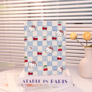 May include: A rectangular tablet case featuring a blue and white checkered pattern with Hello Kitty faces, red bows, and cherries. The case is resting on a stack of books with the text "ATABLE IN PARIS".