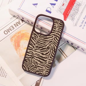 May include: A brown phone case with a zebra stripe pattern. The case has a dark brown border and a tan and brown zebra print design. The phone case is lying on top of a book and newspaper.