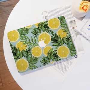 May include: A laptop case featuring a vibrant pattern of yellow lemon slices and green leaves on a light blue background. The case has a white Apple logo in the center. The design evokes a tropical or summery theme.