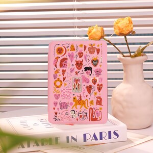 May include: A pink tablet case featuring a colorful illustration of various faces, hearts, and symbols. The case is resting on a book with the title "ATABLE IN PARIS". A white vase with yellow flowers is in the background.