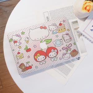 May include: A pink laptop case featuring a repeating pattern of white polka dots and various cartoon characters, including Hello Kitty, cherries, cakes, and stars. The case has a cutout for the Apple logo.