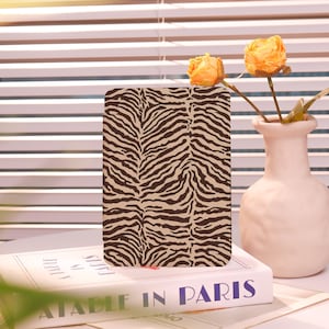 May include: A rectangular case with a zebra stripe pattern in brown and beige. The case is resting on a book titled "A Table in Paris". A small white vase holds two dried orange roses. The background features a window with white blinds.