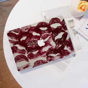 Peut inclure: Une housse d'ordinateur portable avec un motif de baisers de rouge à lèvres rouge foncé sur un fond gris clair. Le logo Apple est visible en blanc. L'ordinateur portable est posé sur une table blanche avec un journal et un livre.