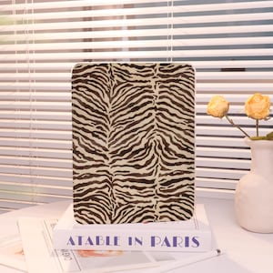 May include: A rectangular tablet case with a zebra print design in shades of brown and cream. The case is resting on a white book with the title "ATABLE IN PARIS" in purple letters. A white vase with yellow flowers is in the background.