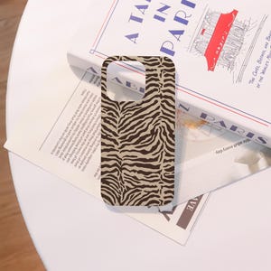 May include: A phone case with a brown and tan zebra stripe pattern. The case is rectangular with cutouts for the camera and buttons. The case is resting on a white surface next to a book titled "A Table in Paris."