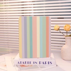 May include: A rectangular case with vertical stripes in pastel colors, including yellow, lavender, green, and peach. The case rests on a book with the text "ATABLE IN PARIS" in navy blue. A white vase with yellow flowers is in the background.