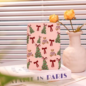 Könnte beinhalten: Eine rosa rechteckige Hülle mit einem sich wiederholenden Muster aus Weihnachtsbäumen, Teddybären und roten Schleifen. Die Hülle liegt auf einem Buch mit den Worten "A TABLE IN PARIS". Eine weiße Vase mit gelben Blumen befindet sich im Hintergrund.