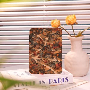 May include: A rectangular, vintage-style print featuring a detailed map of a town in autumn colors. The print is displayed on a book titled "A Table in Paris" next to a white vase with yellow flowers.