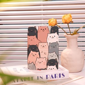 May include: A pink and gray notebook with a cute cat pattern. The cover features a variety of cats with different expressions and poses. The notebook is sitting on top of a book with the title "A Table in Paris".