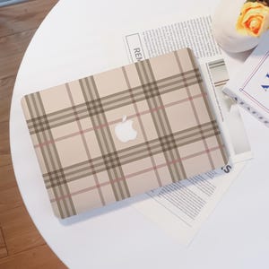 May include: A beige laptop case with a plaid pattern in brown and red. The Apple logo is visible in white. The case is on a white table with a newspaper and a small vase with a yellow flower.