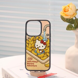 Może przedstawiać: Czarne etui na telefon z kresk&oacute;wkową ilustracją Hello Kitty w zlewie pełnym banan&oacute;w. Projekt zawiera tekst "Hello Hello Kitty's Banana Adventure" w kolorze czerwonym i ż&oacute;łtym.
