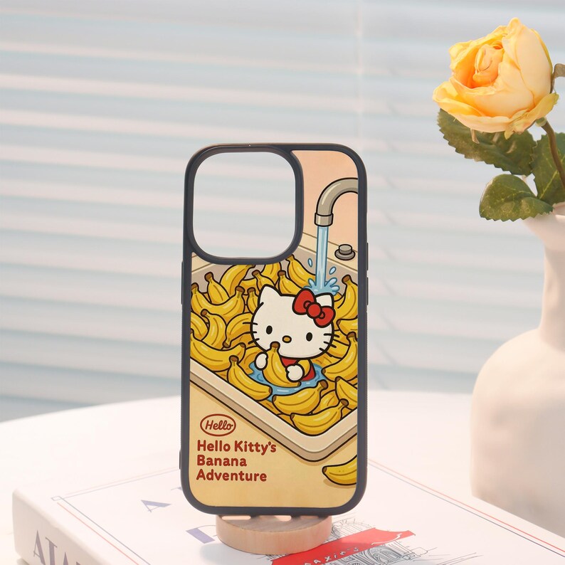 Może przedstawiać: Czarne etui na telefon z kresk&oacute;wkową ilustracją Hello Kitty w zlewie pełnym banan&oacute;w. Projekt zawiera tekst "Hello Hello Kitty's Banana Adventure" w kolorze czerwonym i ż&oacute;łtym.