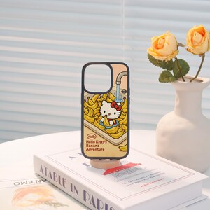 Może przedstawiać: Czarne etui na telefon z motywem Hello Kitty w tematyce banan&oacute;w. Etui ma ż&oacute;łte tło z bananami i kranem wylewającym wodę. Widoczny jest r&oacute;wnież tekst "Hello Kitty's Banana Adventure".