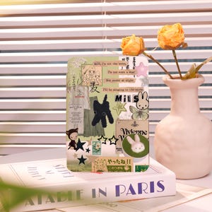 May include: A decorative phone case with a collage design featuring illustrations, text, and a bunny. The case is resting on a book titled "ATABLE IN PARIS" with a vase of yellow flowers in the background.