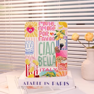 May include: A colorful phone case with an Italian theme. The design includes the words "Ciao Bella" and "Italy", along with illustrations of lemons, sardines, pizza, and other Italian imagery. The case is rectangular and sits on a white book.